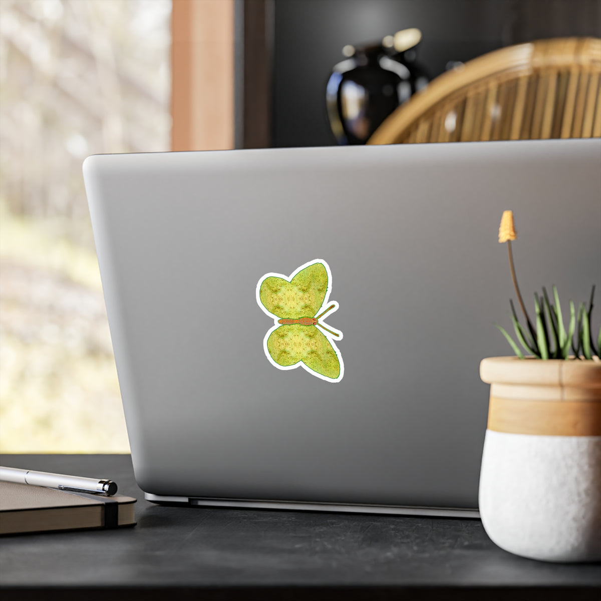 Laptop-lid mock-up: butterfly decal showing pale yellow-green wings patterned with fine olive rosettes, next to a small potted plant.