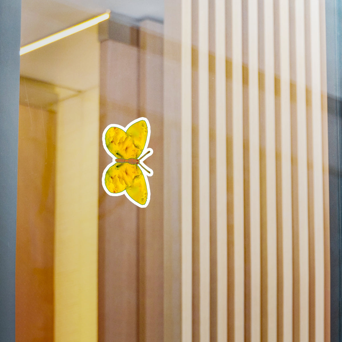 Glass-panel mock-up: white-edged butterfly decal showing golden-yellow wings with soft orange mottling and dark green accents near the body, with pale vertical slats behind.