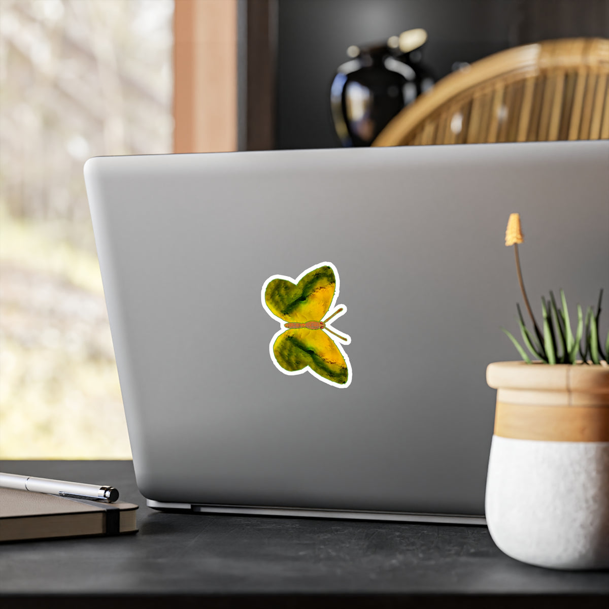 Laptop-lid mock-up: butterfly decal showing yellow wings with broad dark olive bands along the edges, next to a small potted plant.